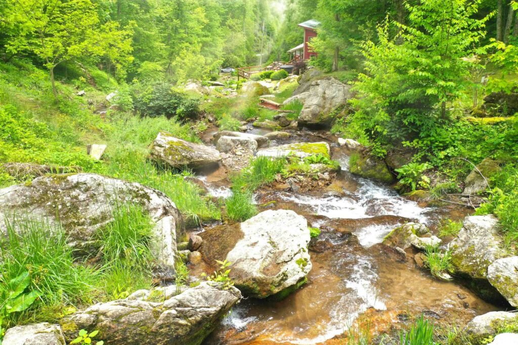 Dronephoto - cascades down to house Secluded waterfall view at Windows Over Waterfalls vacation rental in Hot Springs, North Carolina with mossy rocks, flowing creek water, and forest scenery.